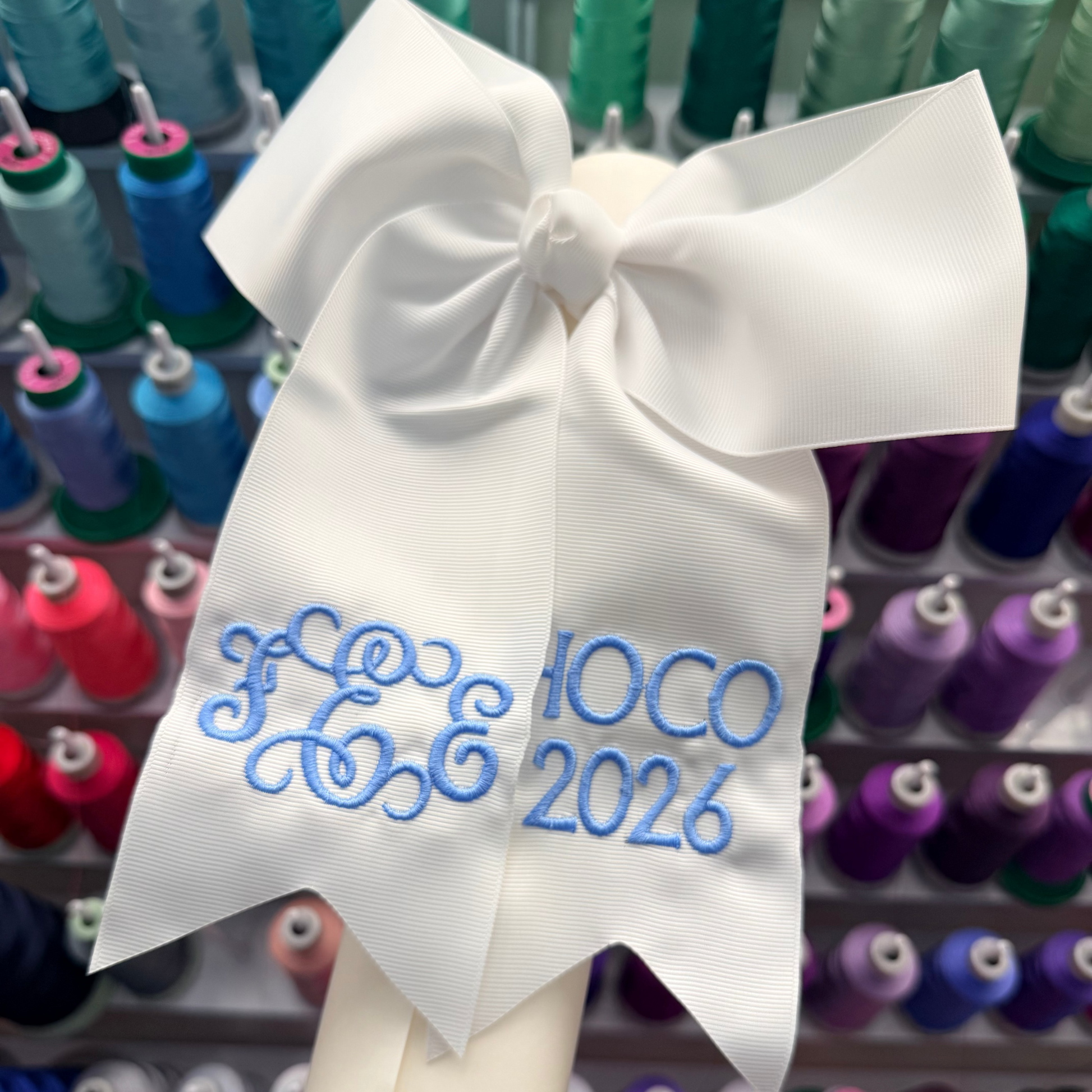 A white fabric hair bow with blue embroidered monogram letters and the text 'HOCO 2026'