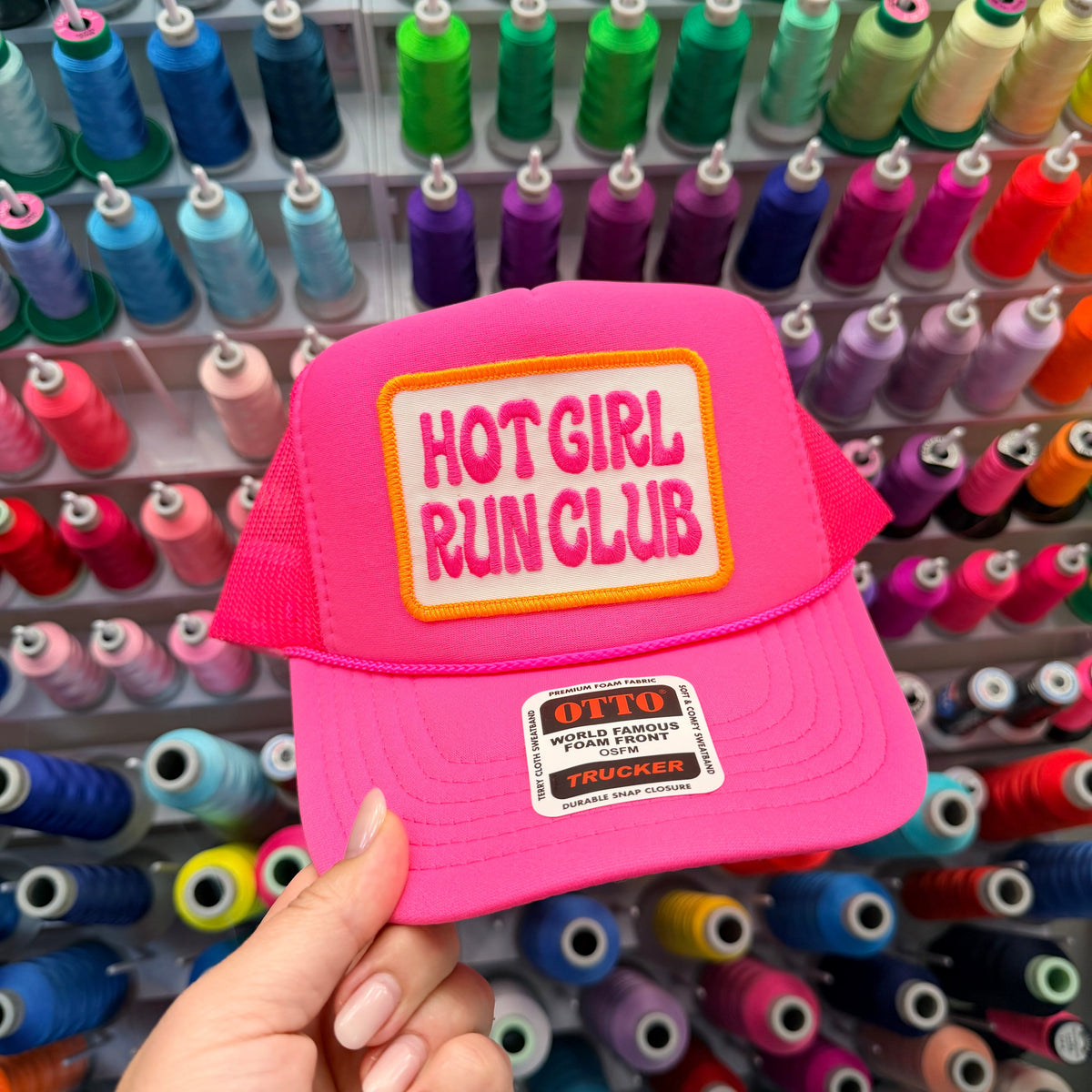 A hand holds a bright pink trucker hat in front of a colorful wall of spools of thread. The hat features a rectangular patch on the front that reads 'HOT GIRL RUN CLUB' in pink letters on a yellow background, and a smaller 'OTTO Trucker' patch on the brim.