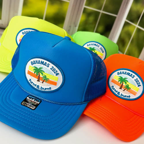Turquoise blue trucker hat with oval palm tree Bahamas 2024 patch, neon hats behind – close-up